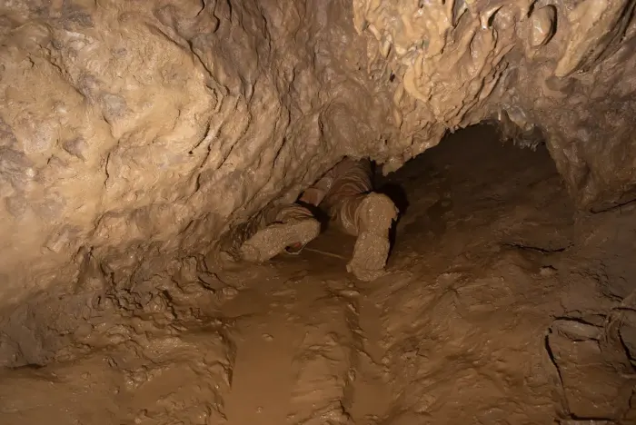A caver crawling through a low, muddy cave passage